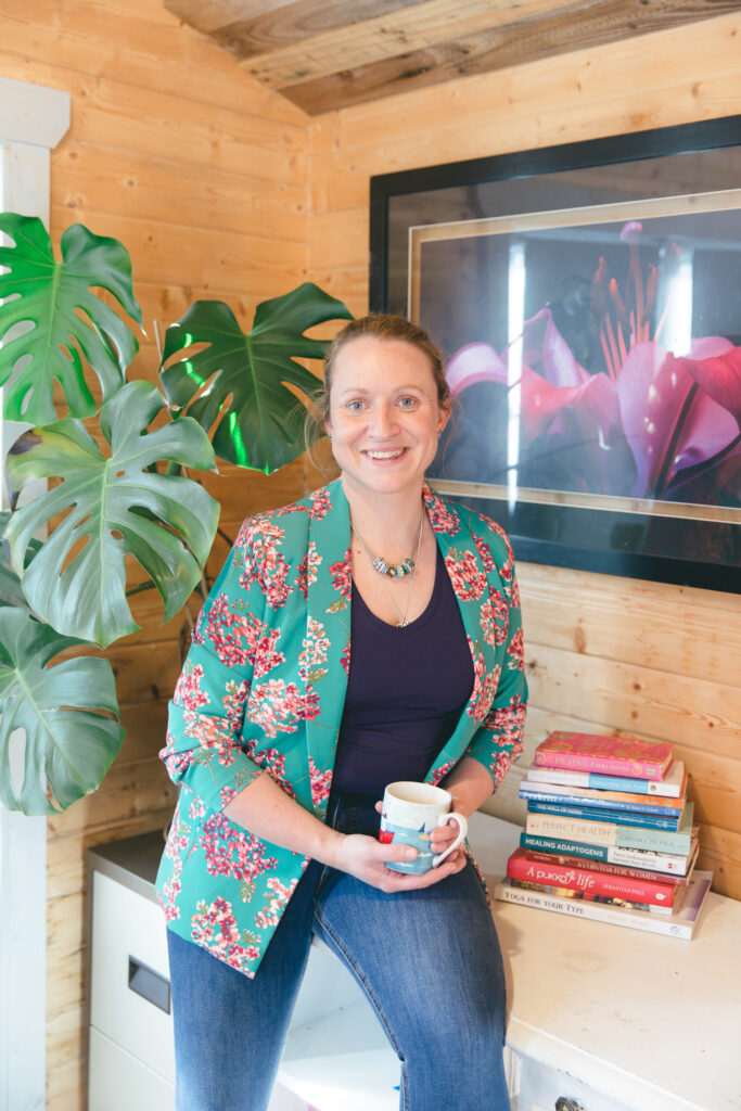 Holly, a white lady with blonde hair, wearing a jade green jacket with pink cherry blossom, jeans and a blue t-shirt, holding a mug in her ayurveda consultation office.